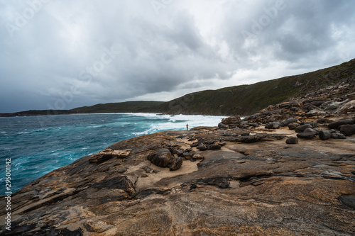 Girl standing at the edge of a cliff watching the rough waves crash against the cliff. Moody sky and overcast weather. Albany  Western Australia. 