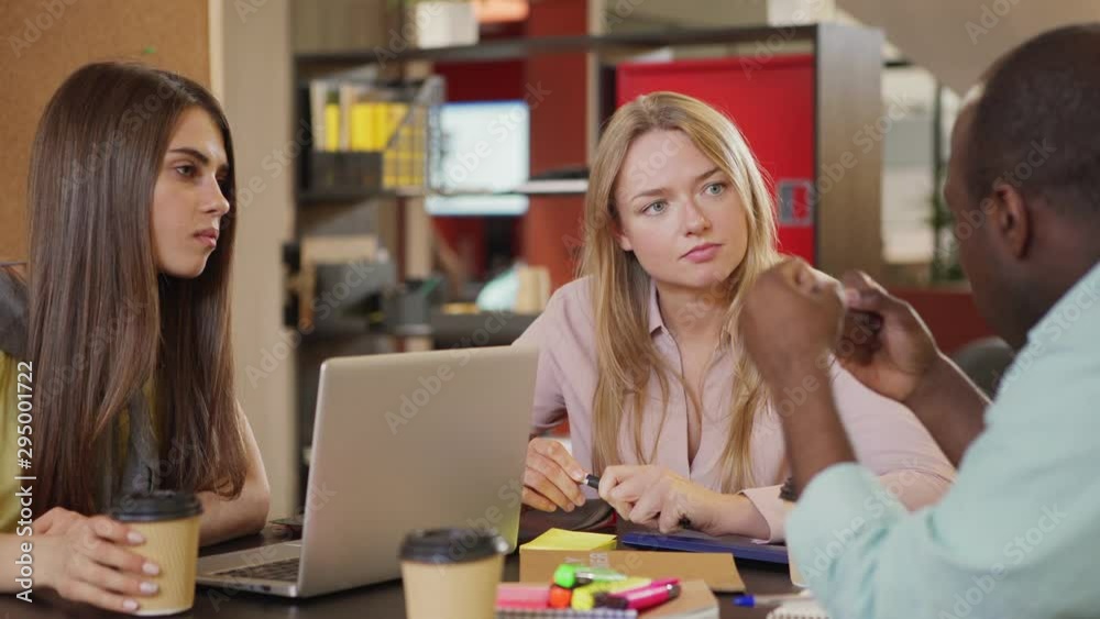 Black man gesturing and talking with female coworkers while sitting at table with laptop and takeaway beverages and working on project together