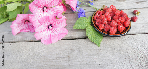 Fototapeta Naklejka Na Ścianę i Meble -  Raspberries in a saucer and flowers on a wooden background. Summer composition with ripe berries. A wide angle image. (Lavatera) flowers. 
