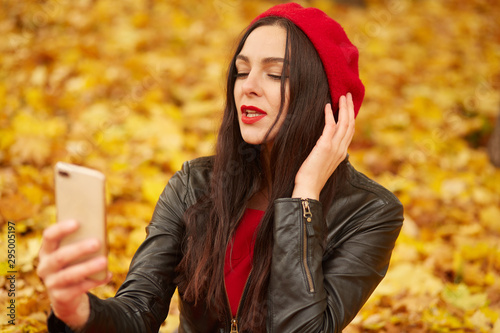 Close up portrait of beautiful young brunette Caucasian woman with her smartphone in hands, taking selfie via phone outdoors in park in autumn, charming lady dresses red beret and leather jacket. photo
