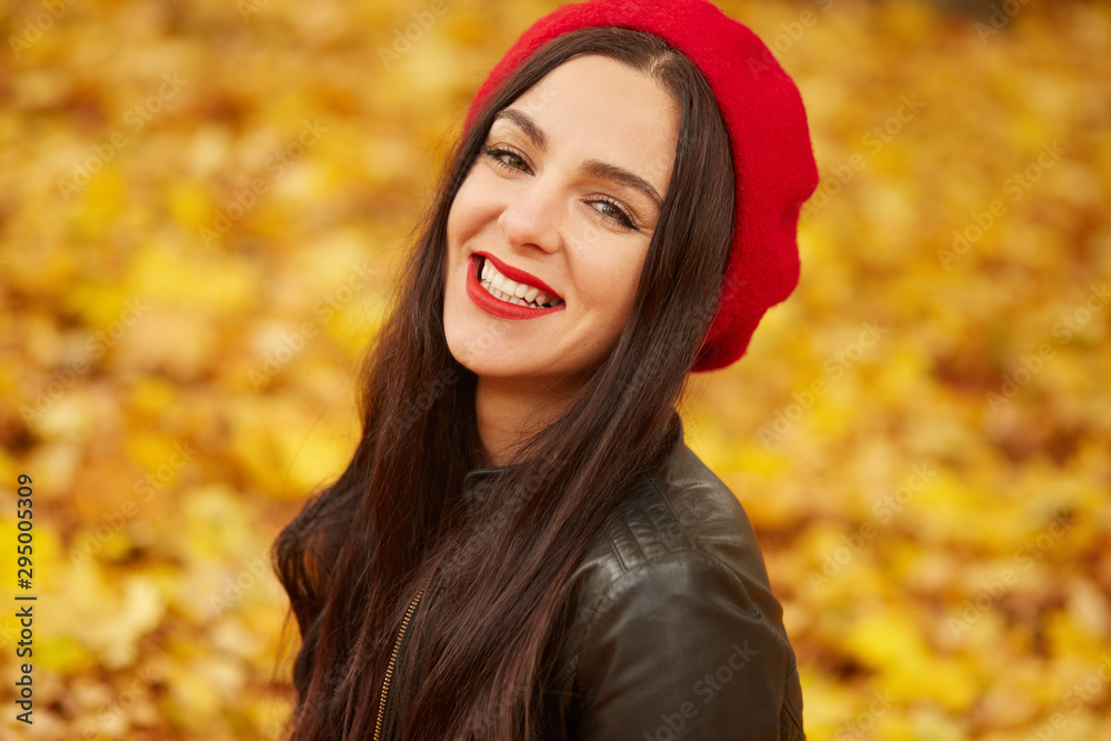 Close up portrait of autumn woman in autumn park, spending sunny days in fall forest, girl wearing red beret and black leather jacket, looking smiling directly at camera. People concept.