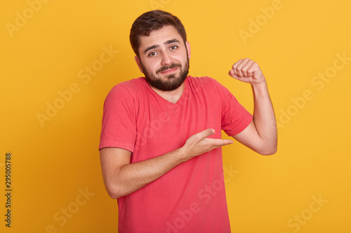 Horizontal shot of male in sleeveless t shirt show his weak biceps muscles, close up portrait of young handsome unshaven man posing isolated over yellow wall background, attractive guy with beard.
