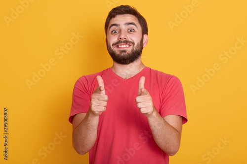 Picture of handsome smiling modern man dresses red casual t shirt showing ok sign with both thumbs, model posing isolated over yellow background, bearded young male with happy facial expression.