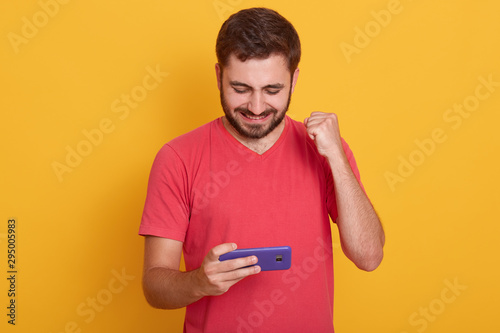 Photo of delighted man dresses red casual t shirt playing video game on cellphone and clenching fist isolated over yellow studio background, bearded guy posing in studio. People and technology. © sementsova321