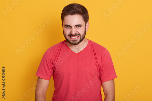 Studio shot of angry man wearing red casual t shirt, being displeased, keeps hands strained, unshaved guy with stylish hairdo twisted his face with anger, posing isolatedover yelow studio background.