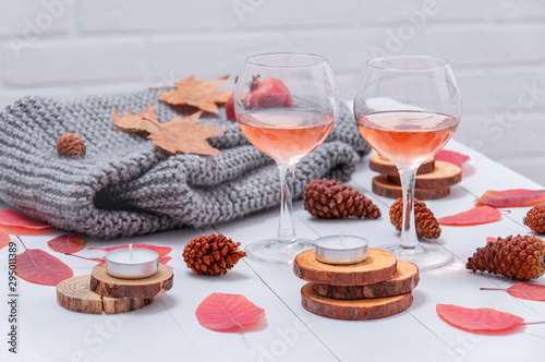 Small candles, two glasses with rose wine, cones, dry red leaves, a gray scarf knitted on a white wooden table. Hello, Autumn. Cozy autumn background.