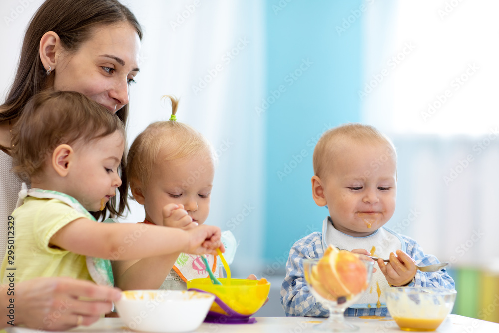 Group of nursery babies eating healthy food with babysitter help. Lunch break in creche. Time to eat in day care