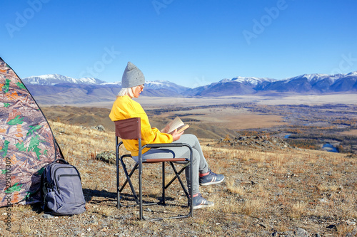 Young woman tourist sitting near the tent and reading a book in the mountains. Travel concept.