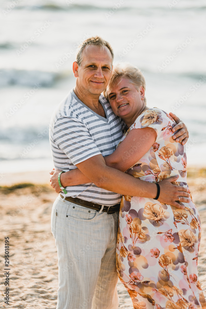 Naklejka premium Romantic adult couple standing together on the beach. Mature pair relax at the seaside on holiday. Happy familiy walking and holding hands near sea. Husband embracing wife. Love story near the sea