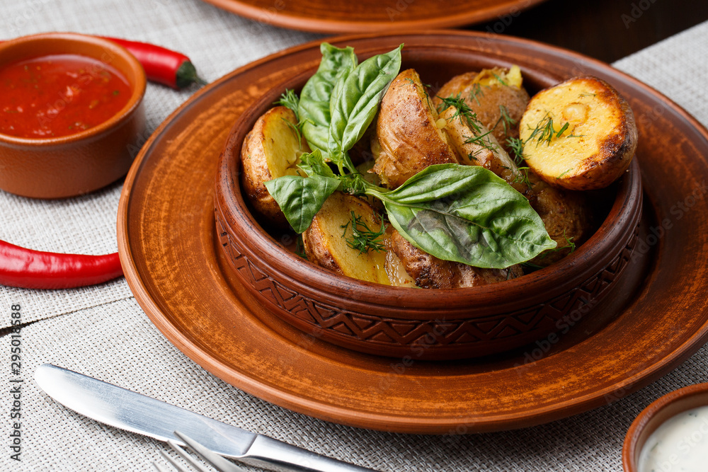 Fried potato in brown clay bowl close-up. Front view of plate with fried potato and basil with sauces and fork with knife. Focus on fried potato. Grey background. horisontal photo