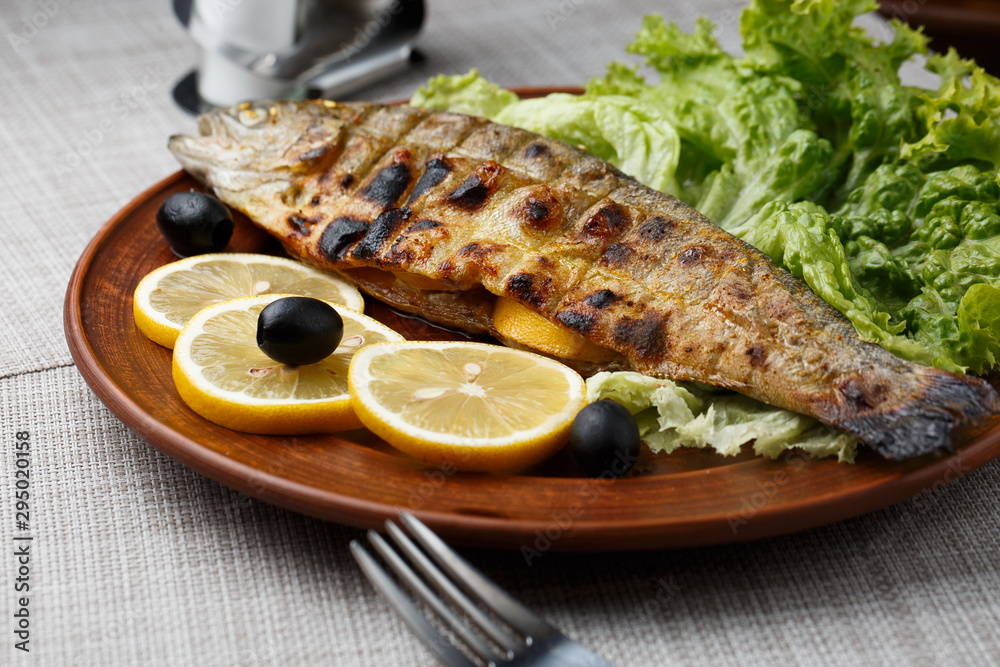 Grilled trout in brown clay bowl close-up. Front view of plate with grilled trout garnishe with salade, lemon and olives. Focus on fried fish. Grey background. horizontal photo