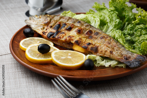Obraz na płótnie Grilled trout in brown clay bowl close-up. Front view of plate with grilled trout garnishe with salade, lemon and olives. Focus on fried fish. Grey background. horizontal photo w salonie