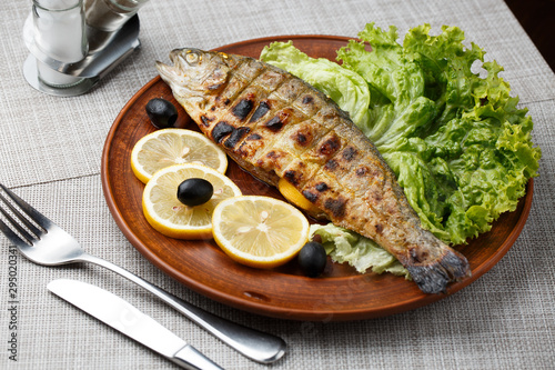Fototapeta Naklejka Na Ścianę i Meble -  Grilled trout in brown clay bowl close-up. Front view of plate with grilled trout garnishe with salade, lemon and olives. Focus on fried fish. Grey background. horizontal photo
