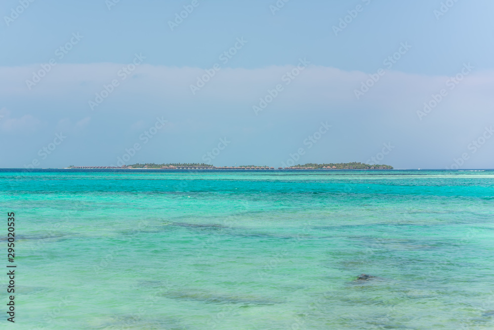 Obraz premium Wide angle picture of the Island on the horizon with bungalows on top of turquoise water in a island located close to Maafushi in Maldives. Picturesque seascape with blue sky.