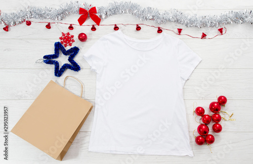Close up white blank template t shirt with copy space paper bag. Christmas Holiday concept. Top view mockup t-shirt, star, white wooden background. Happy New Year decorations accessories. Xmas outfit