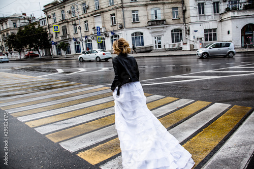 rear view of alone bride running on crosswalk at the intersection. Woman wearing white wedding dress with plume and black jacket