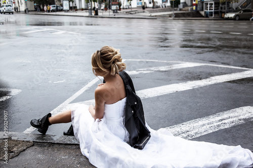 Fotografie Style alone bride sit on a road , elegant model wearing wedding dress and black leather jacket