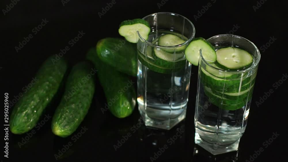 Close-up of a glass of cucumber water on black background, the girl adorns the glass with a cut cucumber. Delicious refreshing water with mint and cucumber in glass. Close-up of a glass of cucumber water on black background, the girl adorns the glass with a cut cucumber. Delicious refreshing water with mint and cucumber in glass.