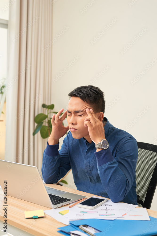 Feeling exhausted. Frustrated young beard man massaging his head and keeping eyes closed while sitting at his working place in office Feeling exhausted. Frustrated young beard man massaging his head and keeping eyes closed while sitting at his working place in office