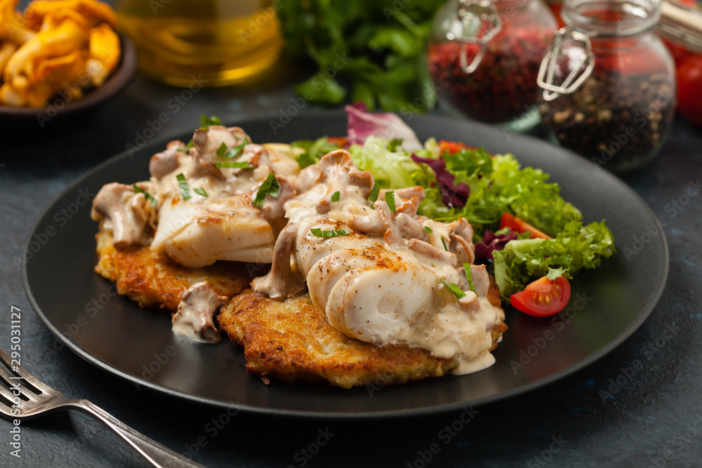 Fried cod, served on potato pancakes with mushrooms sauce and salads. Portion on a black plate. Dark background.