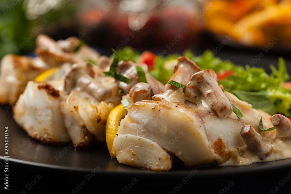 Fried cod served with mushroom sauce and salads. Portion on a black plate. Dark background. Fried cod served with mushroom sauce and salads. Portion on a black plate. Dark background.