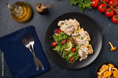 Fried cod served with mushroom sauce and salads. Portion on a black plate. Dark background.