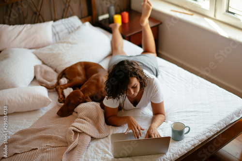 Young woman lying on bed with dog. Girl using laptop while drinking coffee.