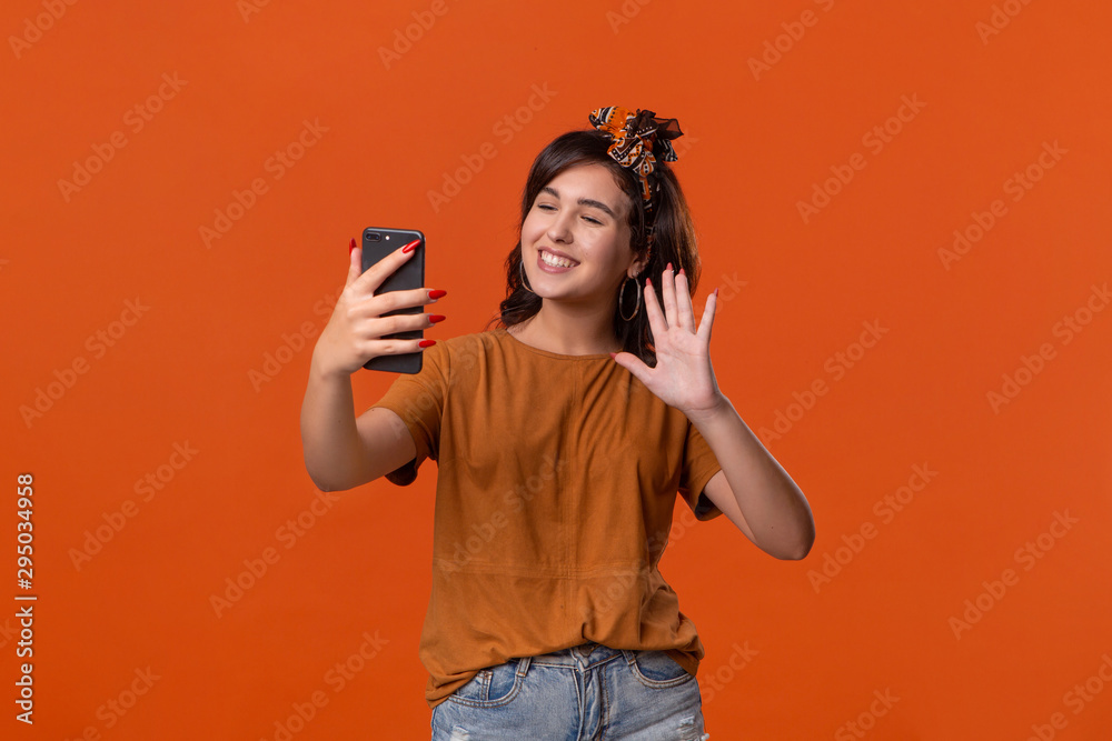 Pretty brunette woman in a t-shirt and beautiful headband making selfie with spartphone standing isolated over orange background. Mobile photography Pretty brunette woman in a t-shirt and beautiful headband making selfie with spartphone standing isolated over orange background. Mobile photography