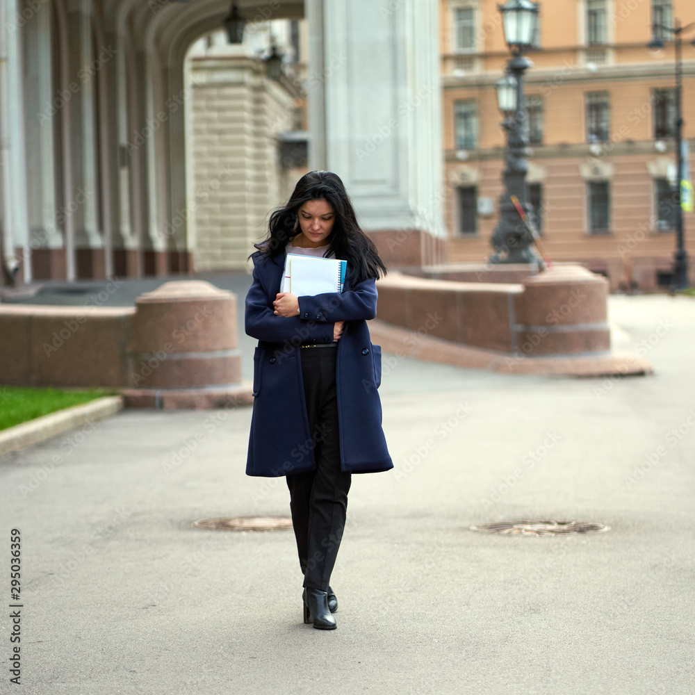 Beautiful serious smart girl brunette student holding notebooks and textbooks, goes walking at University on street of St. Petersburg. A charming woman with long dark hair is studying at course