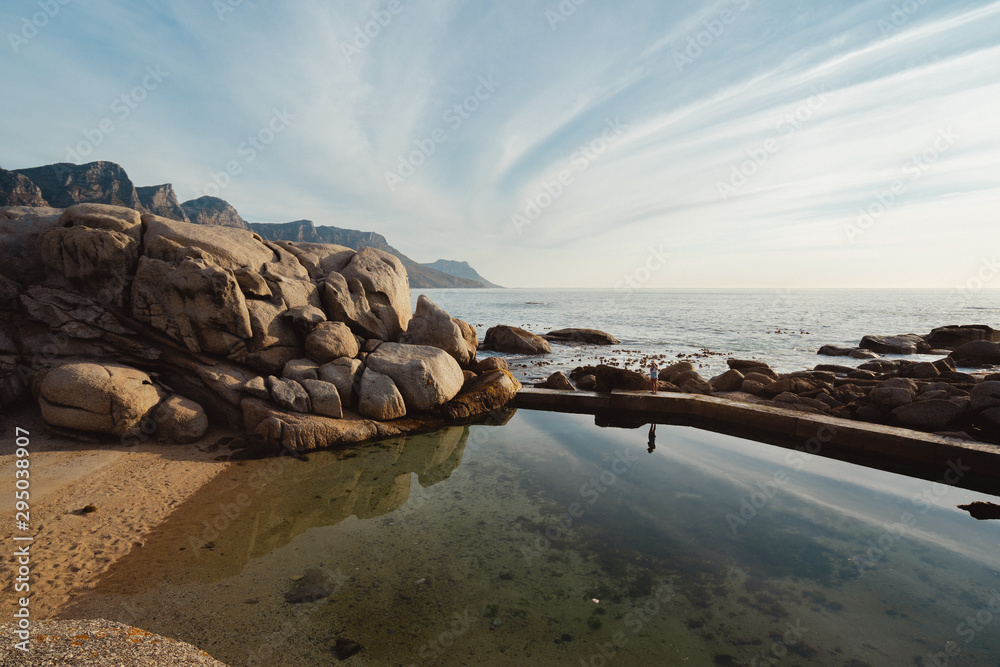 Lone woman standing and reflecting in the pool water, with the ocean and a glimpse of table mountain, a beautiful sunset at Maidens Cove Tidal pool, Camps bay, Cape town, South Africa.