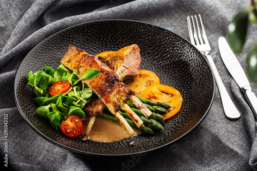 Roasted rack of lamb served with asparagus, yellow tomatoes and lamb sauce. Loin lamb Served in black stone plate with fork and knife. Gray background. Tuna salad. Top view from above horizontal.