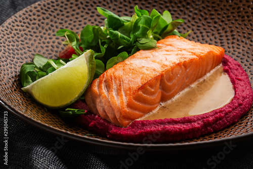 Fried fish salmon fillet with pink humus garnish. One piece of baked salmon grilled lime, tomatoes and salade on a brown plate . Gray background. Horizontal photo.