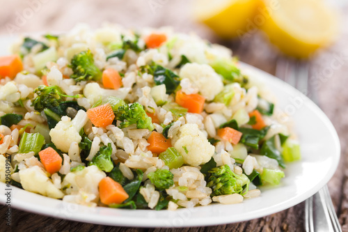 Fresh cooked brown rice with steamed vegetables (broccoli, cauliflower, swiss chard, carrot, celery) on plate, lemon in the back (Selective Focus, Focus one third into the plate)