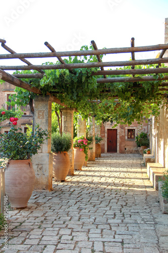 Fototapeta Cozy courtyard of Arkadi monastery with vine trees and flowers in pots