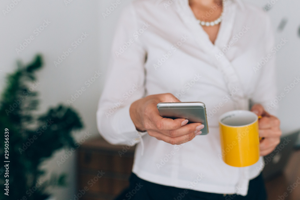 close up of successful businesswoman standing in her office drinking coffee and using mobile phone
