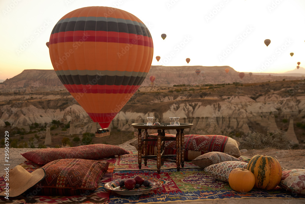Obraz premium big colourful balloon rises in the blue sky above a sandy hill on the ground there is a Turkish carpet with pillows, a plate of fruit and two pumpkins on it and a table in the middle of the carpet
