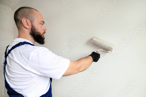 Construction worker doing finish renovation at apartment. Professional painter contractor using white paint roller brush painting of concrete wall room on construction site. Home renovation concept.