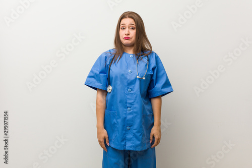 Young nurse woman against a white wall shrugs shoulders and open eyes confused.