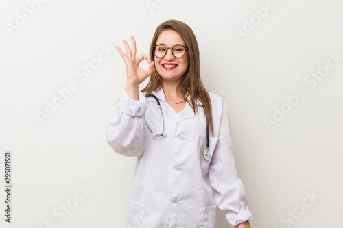Young doctor woman against a white wall cheerful and confident showing ok gesture.