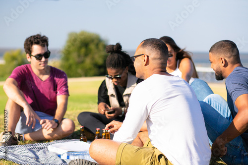 Multiethnic group of friends meeting in park for picnic. Young men and woman sitting around plaid with beer and water bottles. Picnic concept