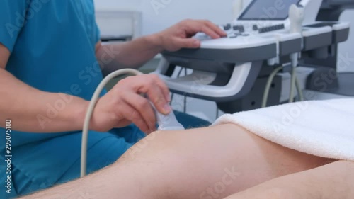 Ultrasound screening of knee joint for man using ultrasound scanner in clinic, closeup view. Doctor runs ultrasound sensor over man's leg, working on scanner panel and looking at screen in hospital.