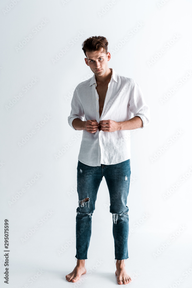 handsome man standing in blue jeans and touching shirt while undressing on white handsome man standing in blue jeans and touching shirt while undressing on white