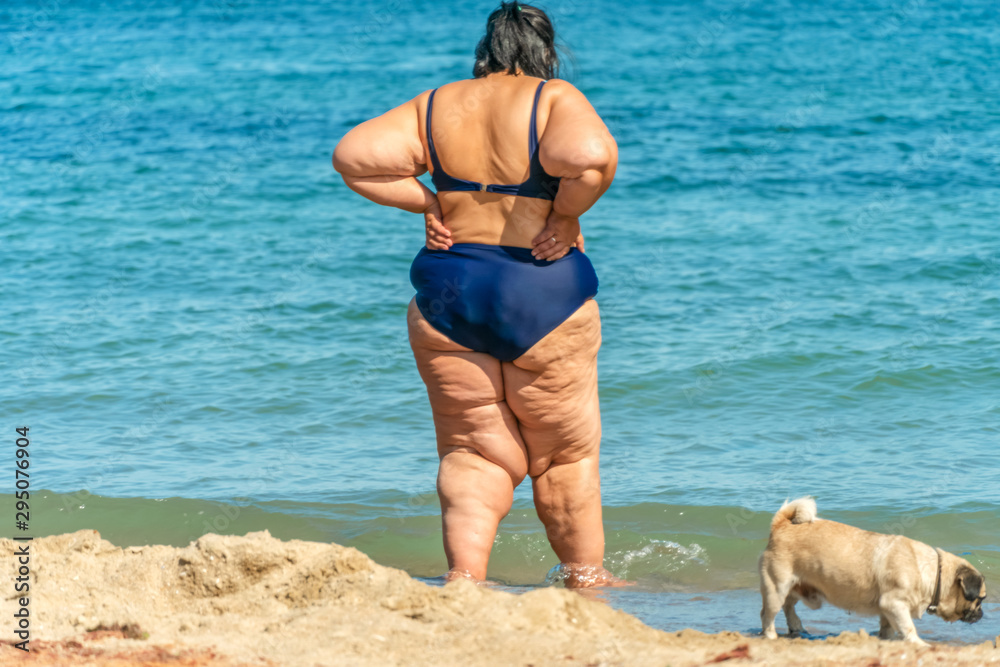 plump woman in a swimsuit stands on the beach with a dog pug and looks into the distance of the sea. Unrecognizable person. photo from the back plump woman in a swimsuit stands on the beach with a dog pug and looks into the distance of the sea. Unrecognizable person. photo from the back