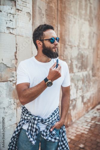 Handsome hipster guy with beard wearing white blank t-shirt and sunglasses smoking e-cigarette