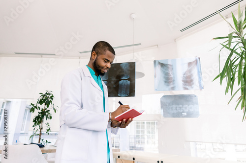 Medicine, oncology, tumour. Professional African male oncologist looking at the X-ray photos of the patients and making notes in red notebook