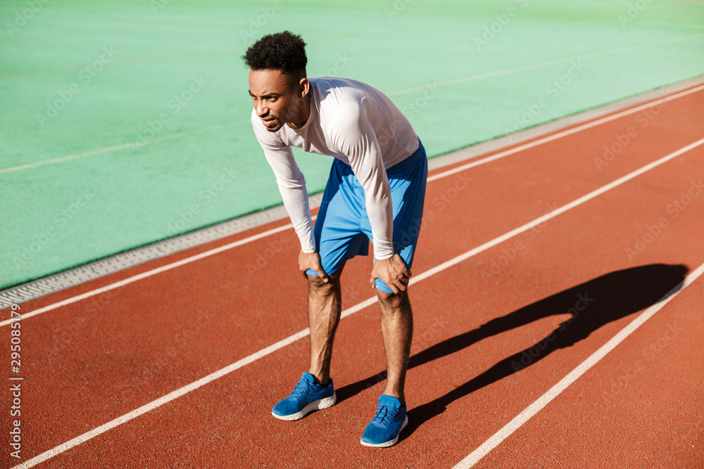 Young tired African American sportsman resting on racetrack after run at stadium