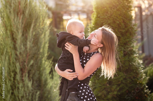 Loving caring young mother kisses her charming one-year-old baby in a black suit on an autumn sunny warm day. Family holiday concept. Smiling happy mom holding little toddler baby in hause garden
