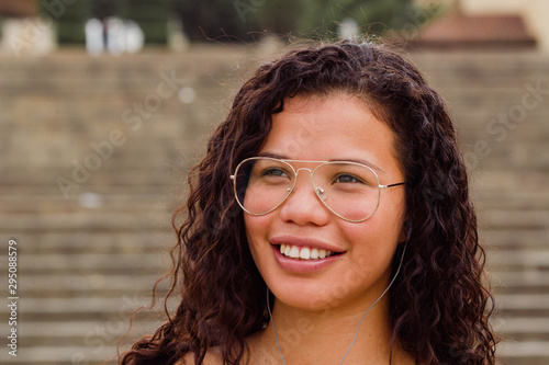 Tablou pe pânză Portrait of a young latin woman listening music and smiling with glasses