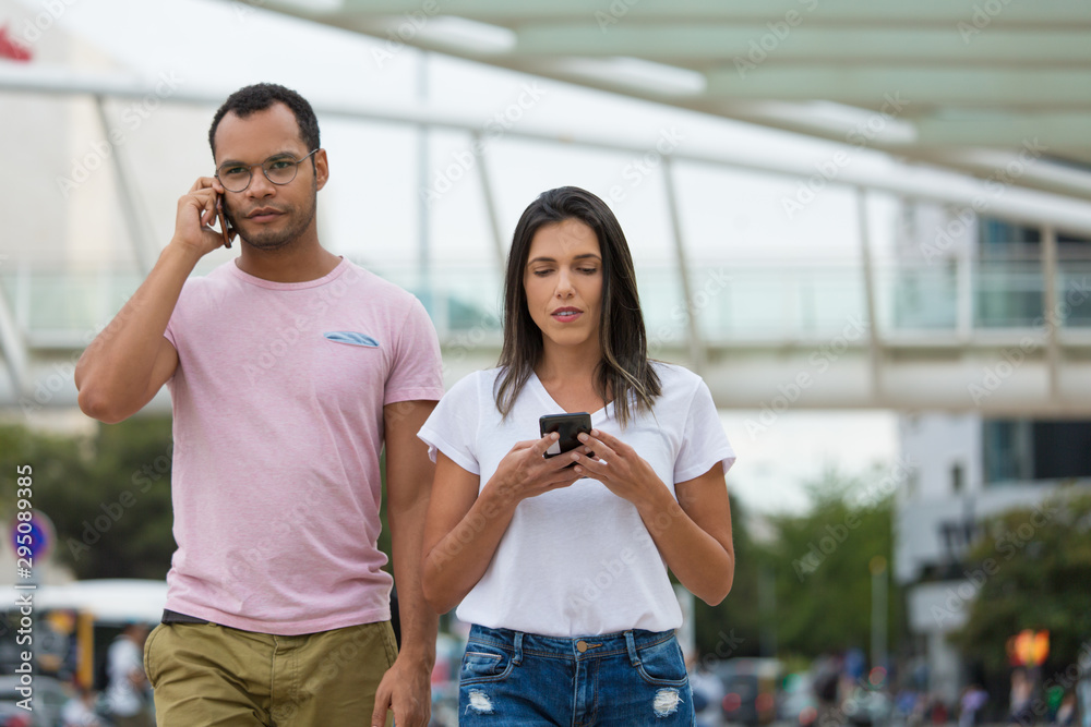Cheerful young people walking on street with smartphones. Smiling African American man talking on phone while Caucasian woman texting nearby. Communication and technology concept