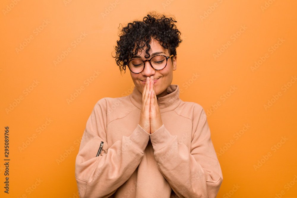 Young mixed african american woman against a brown background holding hands in pray near mouth, feels confident. Young mixed african american woman against a brown background holding hands in pray near mouth, feels confident.